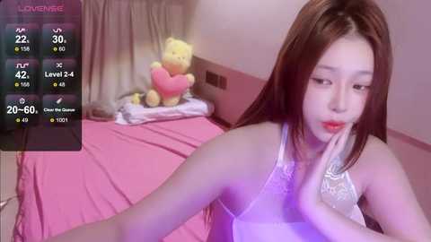 Media: A young Asian woman with long brown hair, fair skin, and red lipstick, wearing a white lace bra, sits on a pink bed with teddy bears.