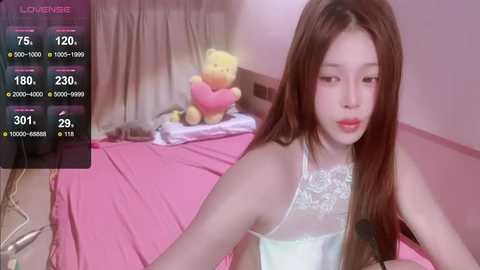 Media: Video of an Asian woman with long brown hair, wearing a white lace lingerie top, sitting on a pink bed with teddy bears.