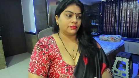 Media: Video of a South Asian woman with medium skin tone, long black hair, wearing a red patterned blouse and black sari with red accents, sitting indoors in a modern, dimly-lit room with wooden furniture, a bed, and a window with blinds.