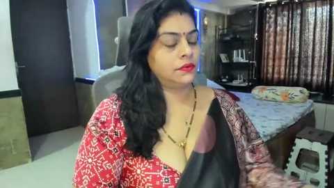 Media: Video of a South Asian woman with medium skin tone and long black hair, wearing a red blouse with black accents, sitting on a gray chair in a modern bedroom with patterned curtains and a bed with floral bedding.