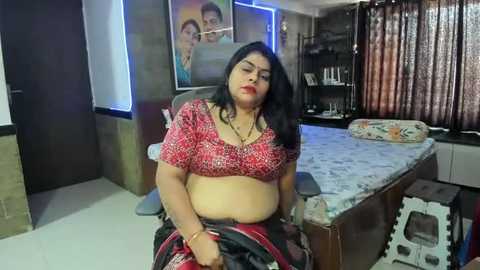 Media: Video of a South Asian woman with long black hair, wearing a red and black patterned blouse, sitting on a bed in a dimly lit room with a poster of a family on the wall.