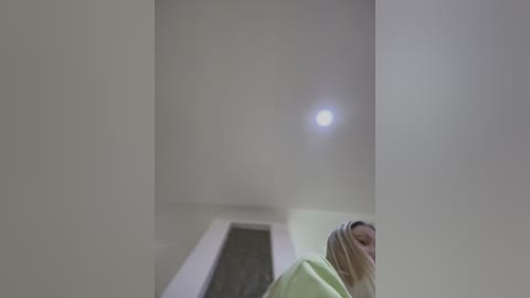 Media: A video of a minimalist, white-walled room with a large, bright light source in the upper right. A blonde woman in a green sweater is partially visible, looking down. The scene has a stark, monochromatic feel.