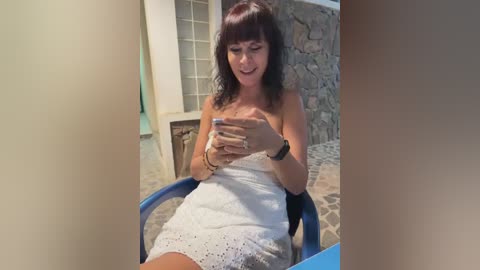 Video of a middle-aged woman with shoulder-length brown hair, wearing a white lace dress, sitting on a blue chair, texting on her phone, in a rustic stone-walled patio.