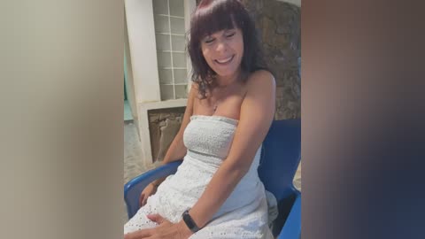 Video of a middle-aged woman with long, dark hair, wearing a strapless white dress, sitting on a blue chair, laughing in a room with stone walls and glass blocks.