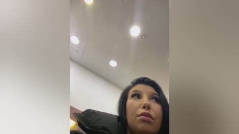 Media: A video of a woman with long black hair and fair skin, wearing a black jacket, looking upward in a room with recessed lights and white walls.