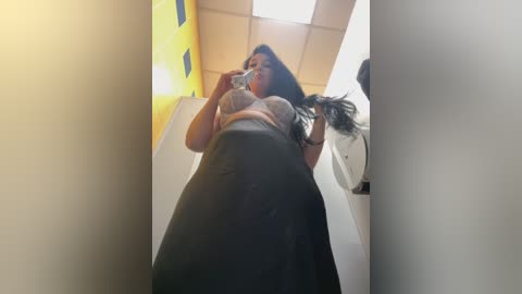 Media: A video of a plus-size woman with long dark hair, wearing a tight gray top and black skirt, taking a selfie in a narrow, brightly lit hallway with yellow and white walls.