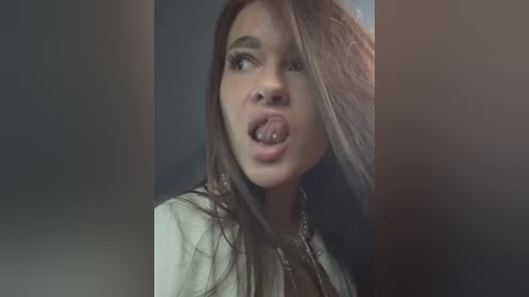 Media: Video of a young woman with long brown hair, pale skin, and a shocked expression, sticking out her tongue, wearing a white shirt. Background is dark and blurry.