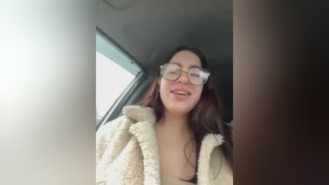 Media: Video of a young woman with light brown skin, long brown hair, wearing glasses and a beige fuzzy robe, smiling inside a car.