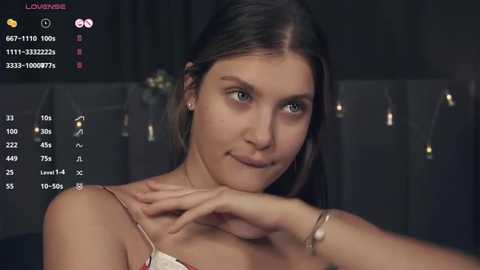 Media: Video of a young woman with light skin and straight brown hair, wearing a beige top. Background shows dimly lit curtains and a recording interface on the left. She appears calm with a slight smile.