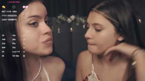 Media: Video of two young women with fair skin, one with long black hair and the other with brown hair, wearing casual outfits, indoors against a dark background.