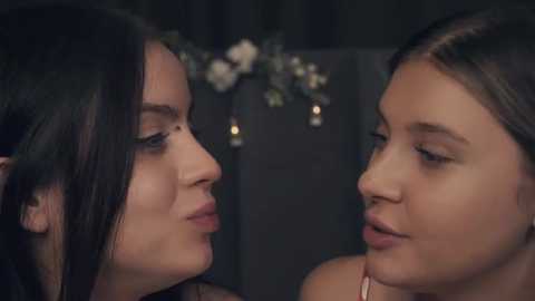 Media: Video of two young women, one with long dark hair and the other with brown hair, gazing at each other with soft, neutral expressions. Background features dim lighting and floral decorations, suggesting an intimate, romantic setting.