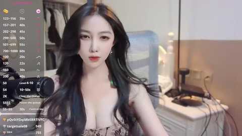 Media: A video of a young Asian woman with long black hair, wearing a low-cut, patterned top, posing in a bedroom with a cluttered desk and a blue fan in the background.