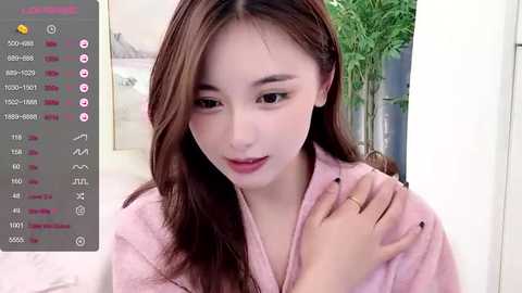 Media: A video of an Asian woman with fair skin, long brown hair, and a pink robe, holding her chin in a thoughtful expression. The background features a green plant and white wall.