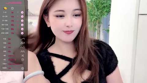 Media: Video of an East Asian woman with fair skin and long, straight brown hair, wearing a black, crisscross-patterned top. Background features a white wall and a green plant.