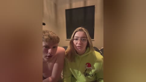 Media: Video of two young women, one topless, sitting on a couch, wearing glasses, smiling, in a dimly lit room with beige walls and a dark window.