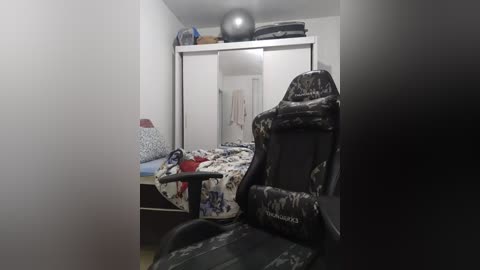 Media: Video of a cluttered, small bedroom with a black gaming chair, white bed, and beige walls. A grey exercise ball and stacked towels atop a shelf.