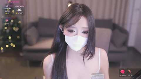 Media: A video of an East Asian woman with long black hair, wearing a white mask, a white spaghetti strap top, and a gold necklace. She stands in a modern, dimly lit living room with a Christmas tree in the background.