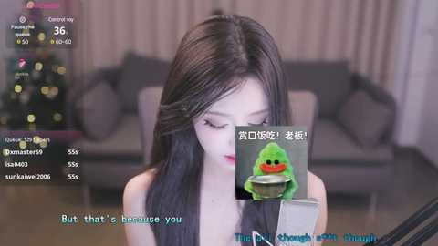 Media: Video of a young East Asian woman with long black hair, wearing a black top, sitting in a living room with a grey couch. A green, smiling emoji with a bowl and spoon is superimposed on the image.