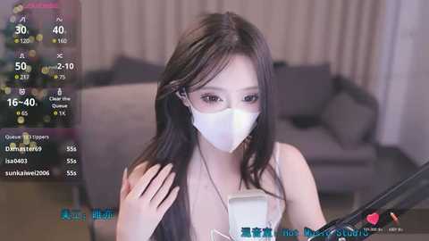 Media: A video of an Asian woman with long black hair, wearing a white mask, in a dimly lit room with a gray couch in the background.