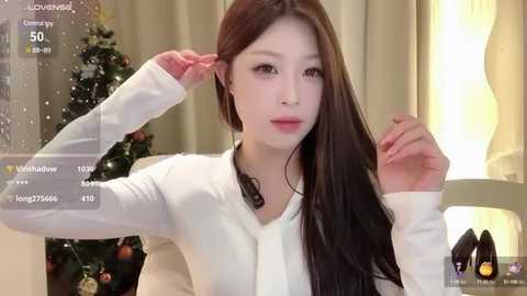 Media: A video of an East Asian woman with long, straight brown hair, fair skin, and a slender physique. She is wearing a white long-sleeve top, adjusting her hair in front of a decorated Christmas tree and a lit lamp in a cozy indoor setting.