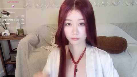 Media: A video of a young woman with long, straight, red hair, fair skin, and wearing a white robe with a red necklace. She's in a cozy bedroom with white walls, a bed, and a wooden nightstand with a candle and books.