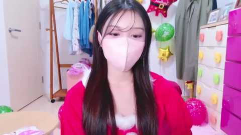 Media: Video of a young East Asian woman with long black hair, wearing a red robe and pink face mask, sitting in a colorful, organized room filled with clothes and toys.