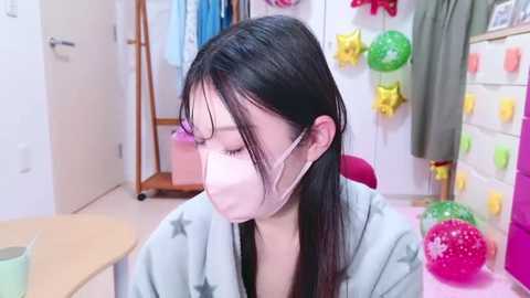 Media: Video of an Asian woman with long black hair, wearing a pink face mask and a light blue robe with star pattern, sitting in a bright, tidy room with white walls, colorful decorations, and a wooden table.