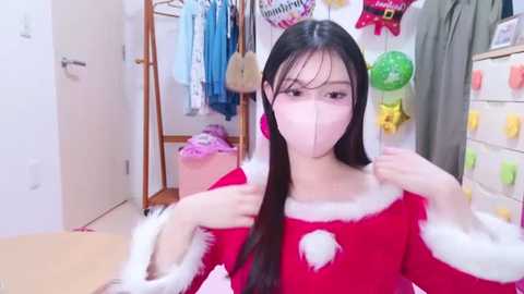 Media: Video of an East Asian woman with long black hair, wearing a pink mask, a red Santa dress with white fur trim, adjusting the dress in a cluttered, brightly lit bedroom with various clothing items hanging.