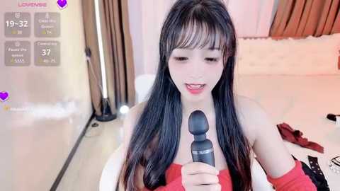 Media: Video of an East Asian woman with long black hair, fair skin, and a slim figure, wearing a strapless red dress. She is holding a black wand vibrator near her lips, smiling. Background shows a white bed with brown curtains and a wooden headboard.