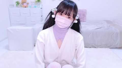 Media: Video of an Asian woman with long black hair, wearing a white bathrobe and a pink face mask, sitting on a white carpet in a minimalist, pastel-colored bedroom.