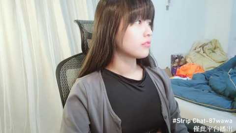 Media: Video of a young Asian girl with long, straight black hair, wearing a black shirt and grey cardigan, sitting on a black chair in a cluttered bedroom with blue sheets and orange clothes.