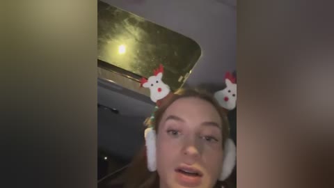 Media: Video of a young woman with fair skin and long brown hair, wearing white headphones with red and white rooster ears. She's in a dimly lit room, looking into a reflective surface.