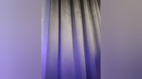 Media: Video of a pair of long, grey curtains hanging against a plain, light grey wall. The curtains have a smooth, slightly textured fabric and are slightly draped, with a faint purple glow at the bottom left corner.