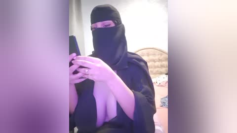 Media: Video of a woman in a black niqab, holding a smartphone, in a bedroom with a bed and pillows in the background.