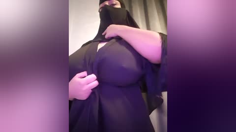 Media: Video of a person with a light brown complexion, wearing a black hijab, partially lifting it to reveal a large, round breast. The background is blurred with muted purple and pink tones.