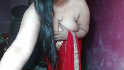 Media: Video of a topless woman with long black hair, wearing a red sari with gold trim, partially covering her breasts with her hand, in a dimly lit room with a purple wall and indistinct background.