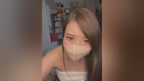 Media: Video of a young Asian woman with long brown hair, wearing a white mask, standing in a cluttered room filled with toys and books, looking directly at the camera.