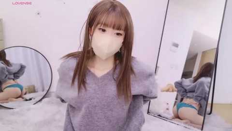 Media: Video of an East Asian woman with long, straight brown hair, wearing a gray sweater, teal panties, and a white mask, kneeling in front of a mirror, reflected image showing her buttocks.