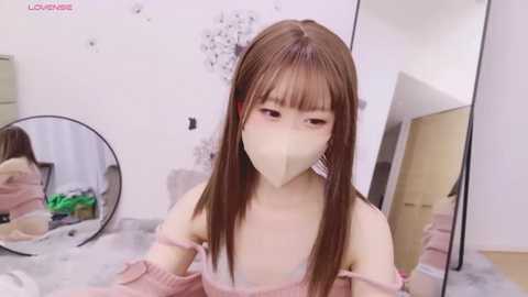 Media: Video of a young Asian woman with straight brown hair, wearing a beige face mask, pink off-shoulder sweater, and sitting on a bed with a round mirror reflecting her reflection.