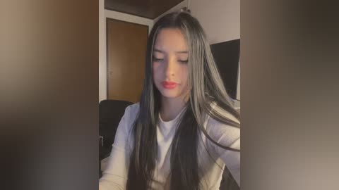 Media: Video of a young Asian woman with long black hair, fair skin, and red lipstick, wearing a white sweater, seated indoors with a blurred background.