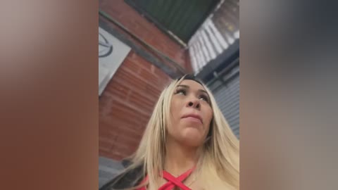 Media: Video of a blonde woman with straight hair, wearing a red top, looking up, taken from a low angle, with blurred background of a brick wall and metal gate.