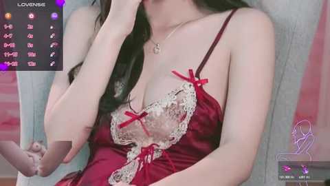 Media: Video of a fair-skinned woman with long dark hair, wearing a revealing red satin lingerie with lace trim and red bows. She's seated, partially covering her face with her hand, in a softly lit room.