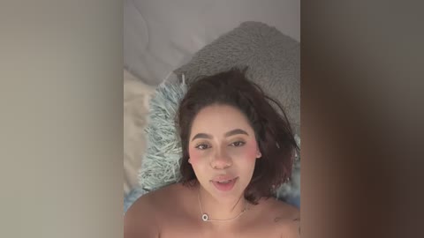 Media: Video of a young, light-skinned woman with dark hair lying on a bed, wearing a necklace. She has a small, round belly and is topless, with a blurred background featuring soft pillows.