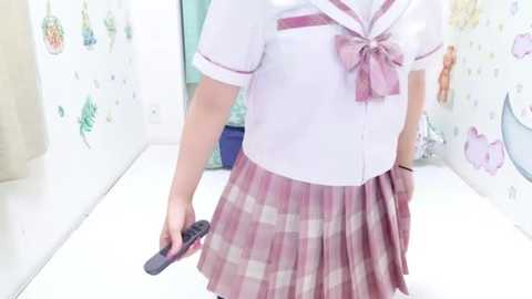 Media: Video of a petite, fair-skinned woman wearing a white sailor schoolgirl outfit with a pink plaid skirt and bow, holding a black cell phone, standing in a brightly lit, minimalistic room with white walls and pastel decor.