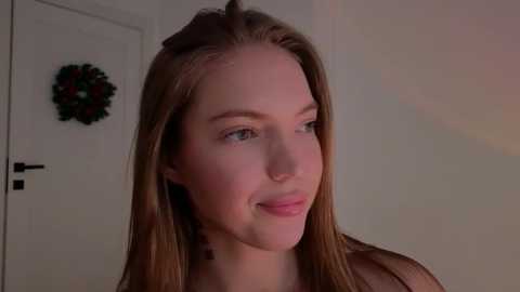 Media: Video of a fair-skinned young woman with long, straight, light brown hair, wearing a sheer top, standing indoors next to a white door adorned with a festive wreath.