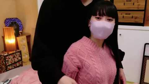 Media: Video of a woman in a pink knit sweater and white mask, standing close to a man in a black shirt, in a cozy room with wooden furniture and warm lighting.