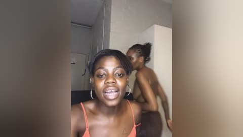 Media: Video of a dark-skinned woman with short hair wearing an orange tank top, standing in a beige-tiled room, looking at her reflection in a mirror.