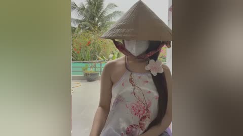 Media: Video of a woman in traditional Vietnamese attire, including a conical hat, floral-patterned top, and mask, standing outdoors amidst lush tropical plants.