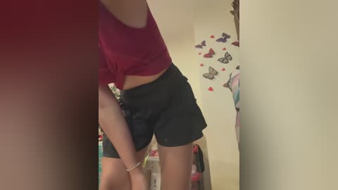 Media: A video of a young woman with medium skin tone, wearing a pink crop top and black shorts, bending slightly, in a cluttered room with butterfly decals and hanging clothes.