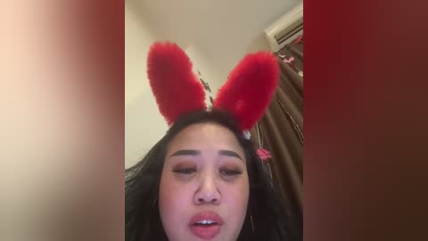 Media: Video of a young woman with long black hair, wearing red bunny ears and dark makeup, against a blurred indoor background.